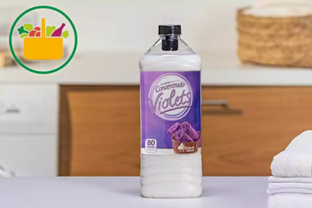 Botella de suavizante concentrado Violets sobre una mesa blanca en una lavandería con toallas dobladas y un ícono de canasta de compras en la esquina superior izquierda Botella de suavizante concentrado Violets sobre una mesa blanca en una lavandería con toallas dobladas y un ícono de canasta de compras en la esquina superior izquierda