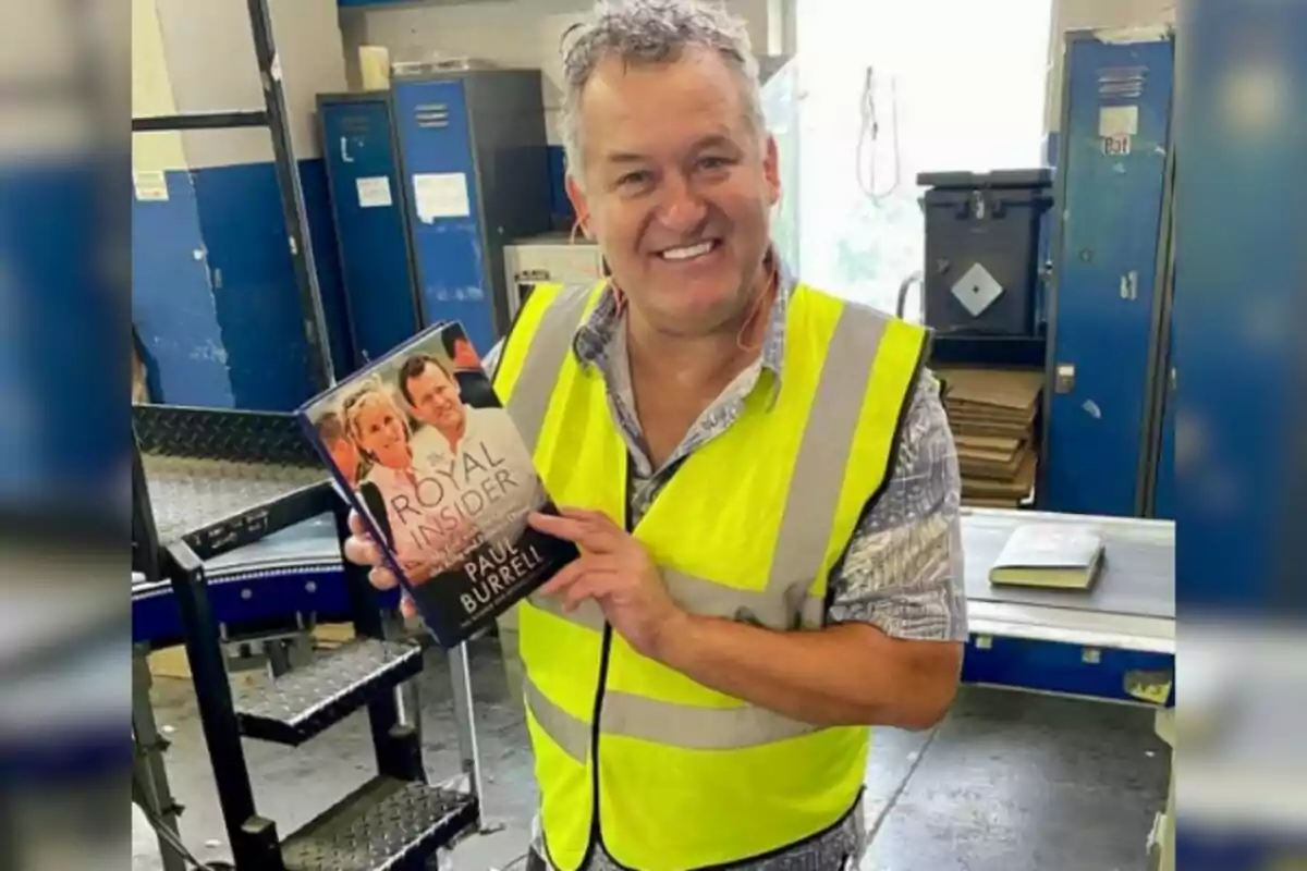 Hombre sonriente con chaleco amarillo sostiene un libro titulado Royal Insider en un entorno industrial Hombre sonriente con chaleco amarillo sostiene un libro titulado Royal Insider en un entorno industrial