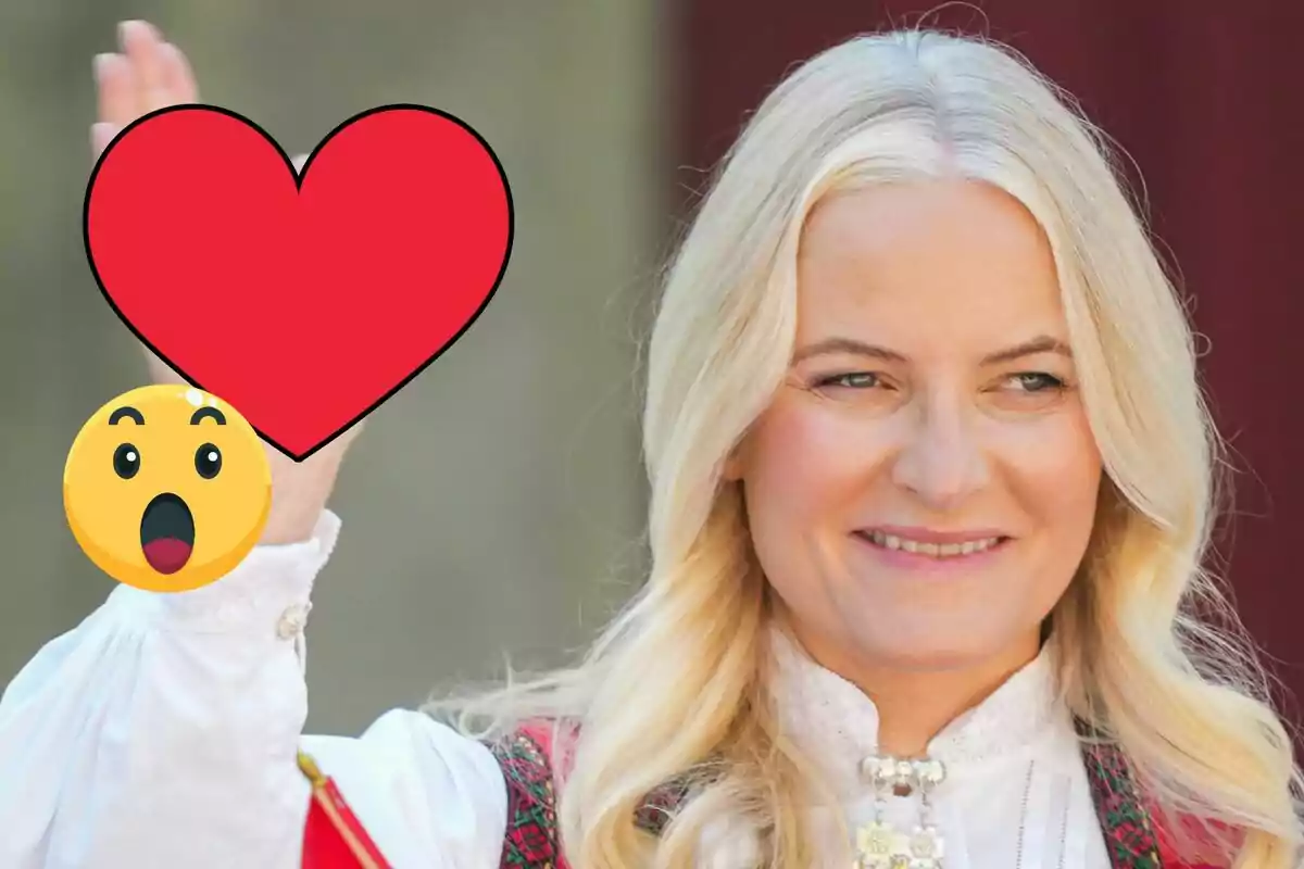 Mette-Marit sonriente con blusa blanca y chaleco tradicional, levantando la mano decorada con un emoji de corazón rojo y una carita sorprendida