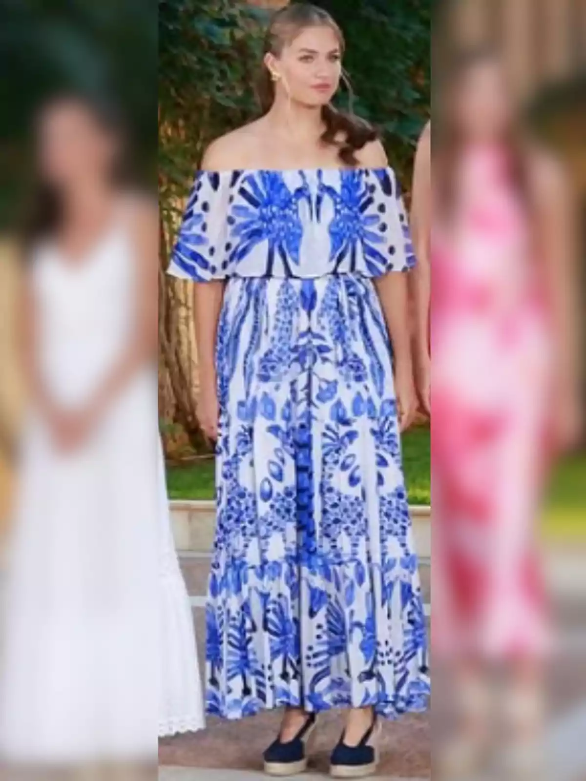 Joven de pie con vestido largo azul y blanco de hombros descubiertos y sandalias oscuras, fondo de vegetación y personas desenfocadas a los lados Joven de pie con vestido largo azul y blanco de hombros descubiertos y sandalias oscuras, fondo de vegetación y personas desenfocadas a los lados