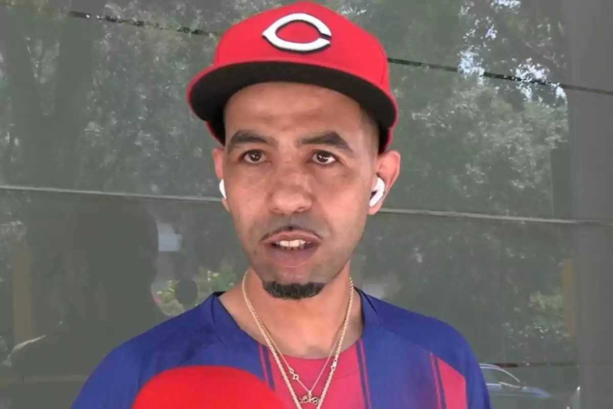 Mounir Nasraoui wearing a red cap and a blue and red t-jersey, using white headphones and gold chains while speaking in front of a red microphone. Mounir Nasraoui wearing a red cap and a blue and red t-jersey, using white headphones and gold chains while speaking in front of a red microphone.