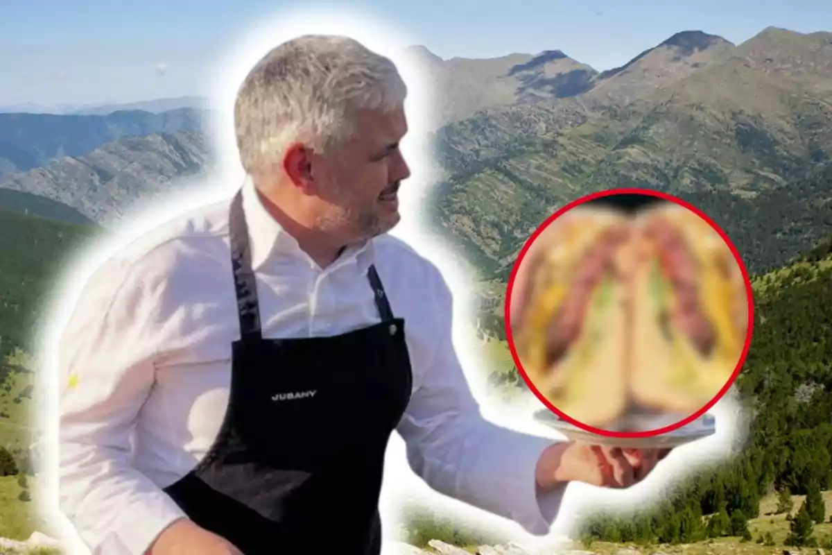 Xef amb cabells canosos i davantal negre sosté un plat amb menjar desenfocat en un entorn muntanyós.