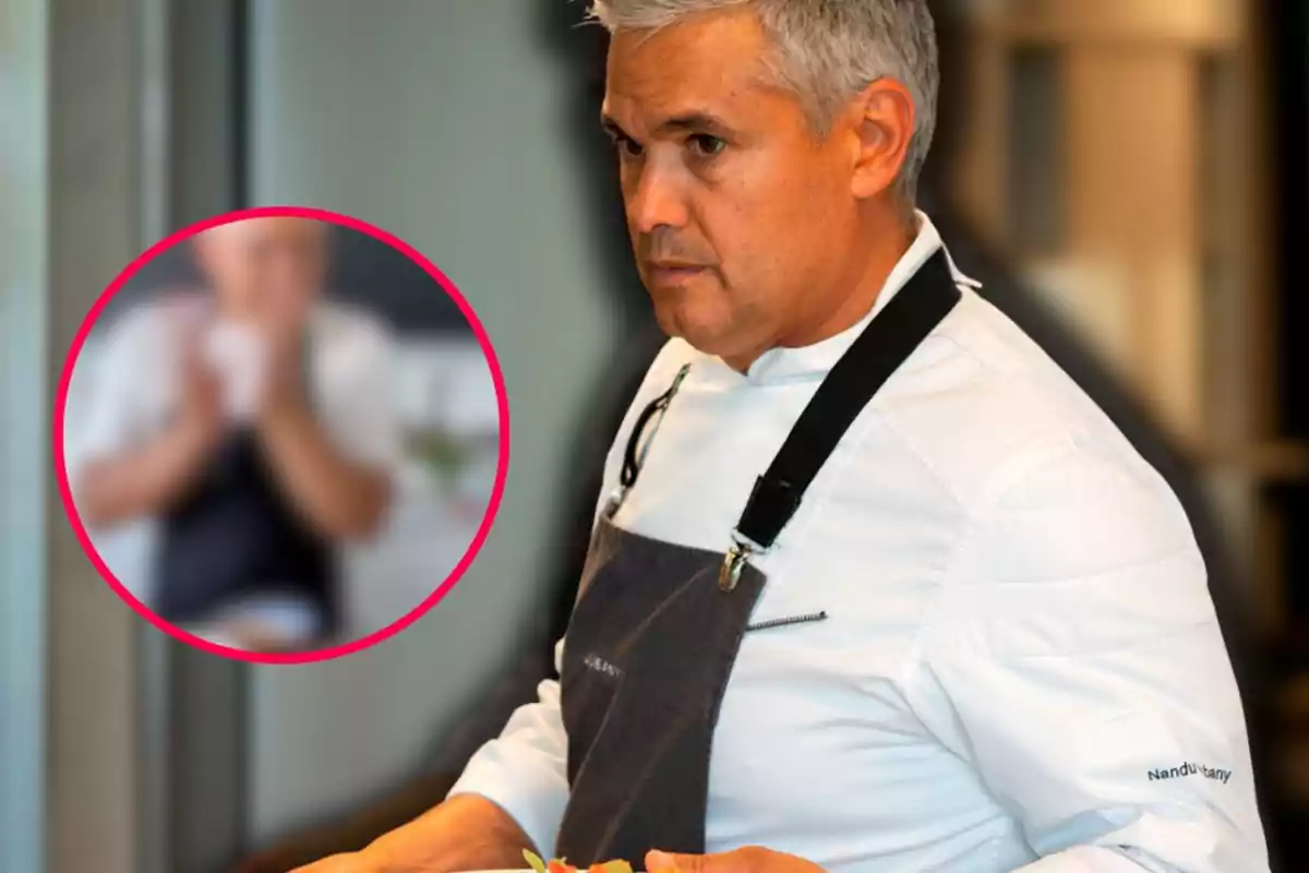 Chef de cabello canoso con chaqueta blanca y delantal oscuro sosteniendo un plato mientras mira hacia el frente con expresión seria y al fondo se observa una figura desenfocada dentro de un círculo rosa.