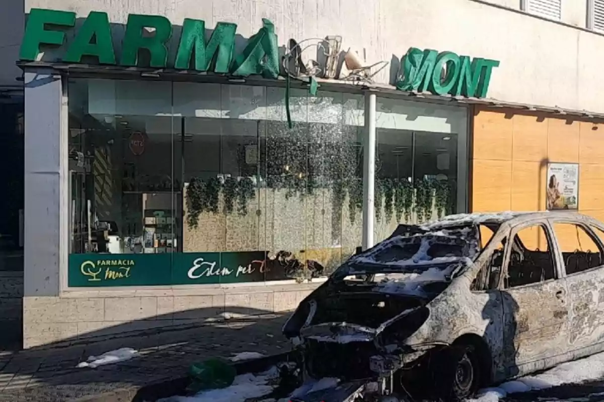 Un coche completamente calcinado está estacionado frente a una farmacia con el letrero dañado y el cristal roto