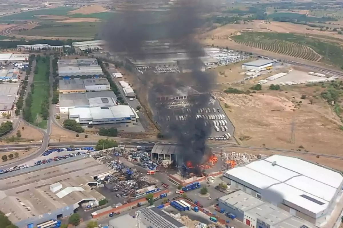Vista aérea de un incendio en una zona industrial con una gran columna de humo negro elevándose sobre los edificios y llamas visibles en una nave industrial rodeada de almacenes y vehículos