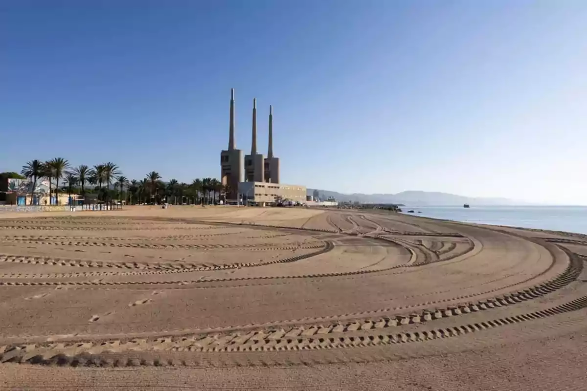 Playa de arena con huellas de llantas y una central eléctrica de tres chimeneas al fondo junto al mar en un día despejado Playa de arena con huellas de llantas y una central eléctrica de tres chimeneas al fondo junto al mar en un día despejado