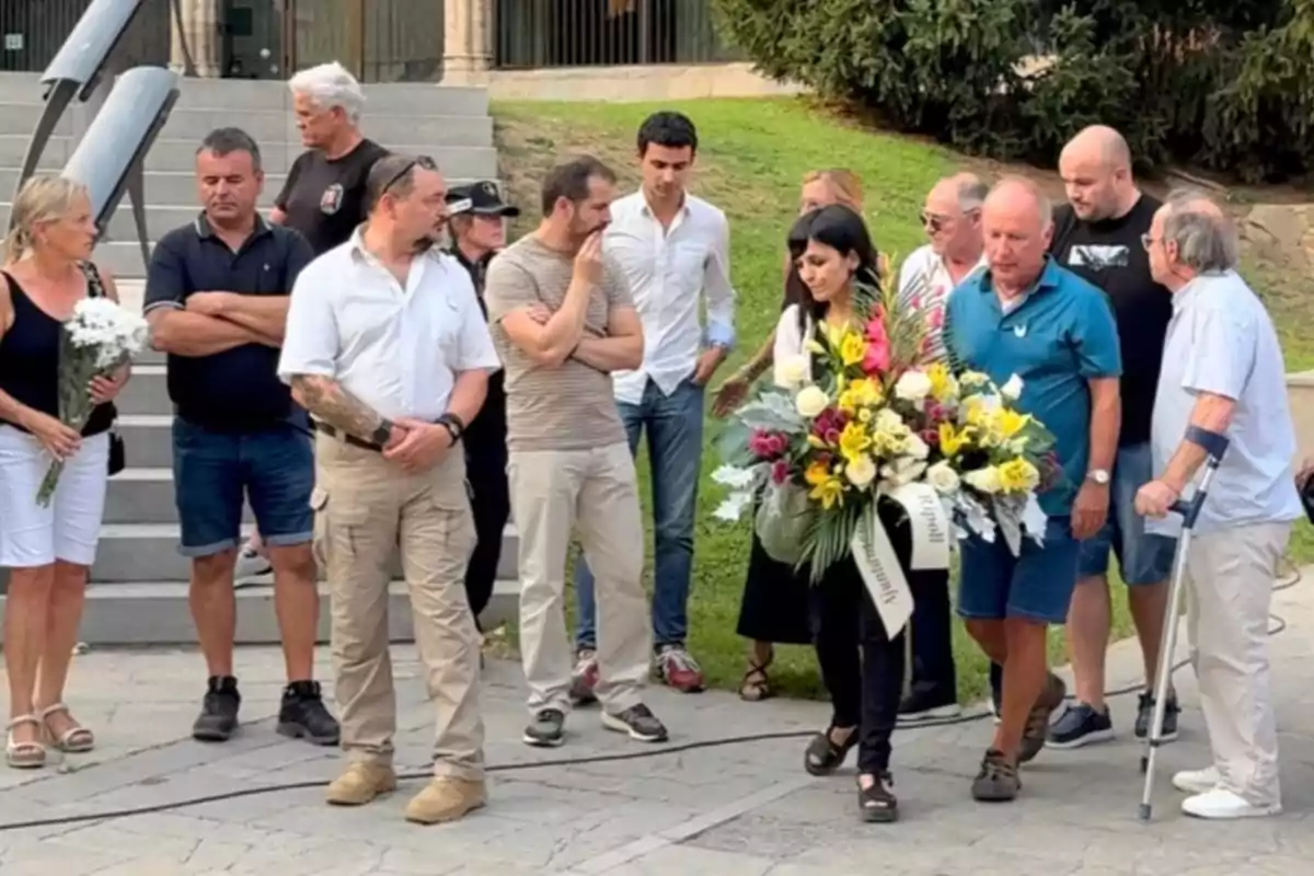 Un grup de persones està reunit a l’aire lliure, algunes sostenen rams de flors i una dona porta un gran arranjament floral, tots semblen participar en un acte commemoratiu o d’homenatge.