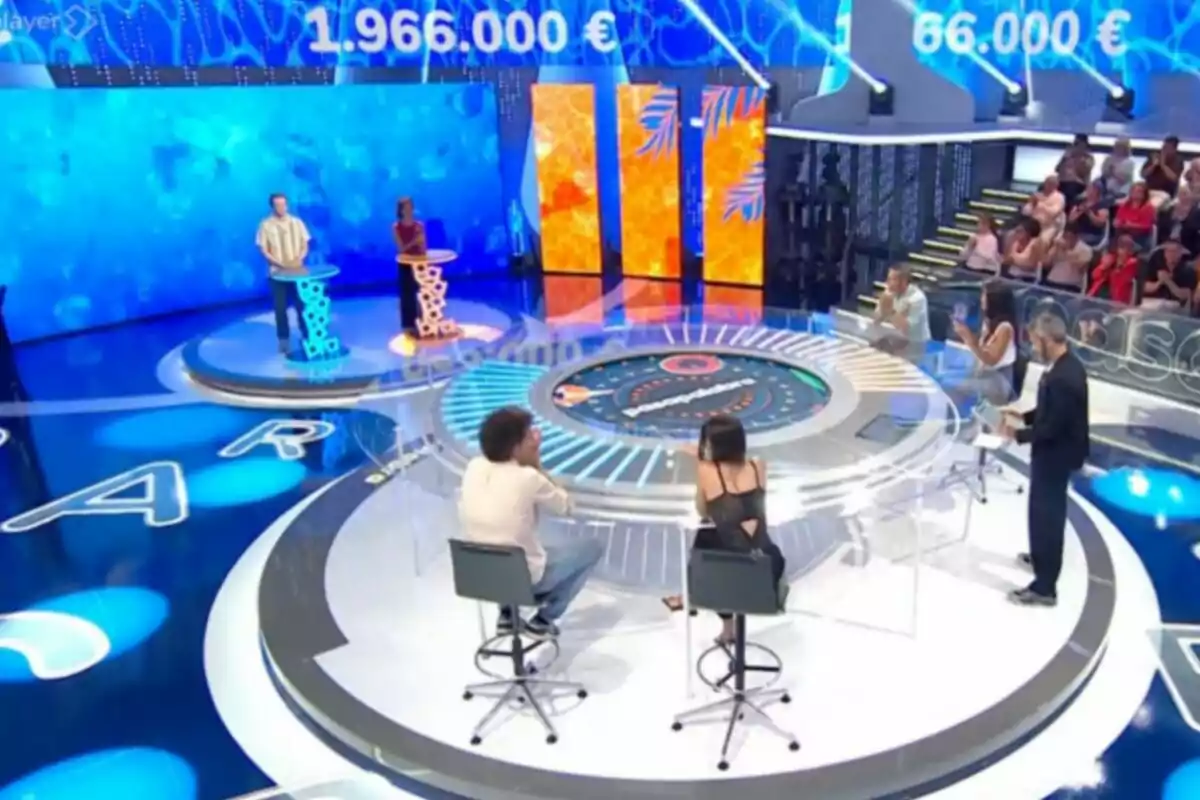 Un plató de concurs televisiu amb participants drets als faristols, un presentador i públic aplaudint a les grades, amb pantalles digitals mostrant xifres grans en euros. Captura de 'Pasapalabra' Un plató de concurs televisiu amb participants drets als faristols, un presentador i públic aplaudint a les grades, amb pantalles digitals mostrant xifres grans en euros. Captura de 'Pasapalabra'