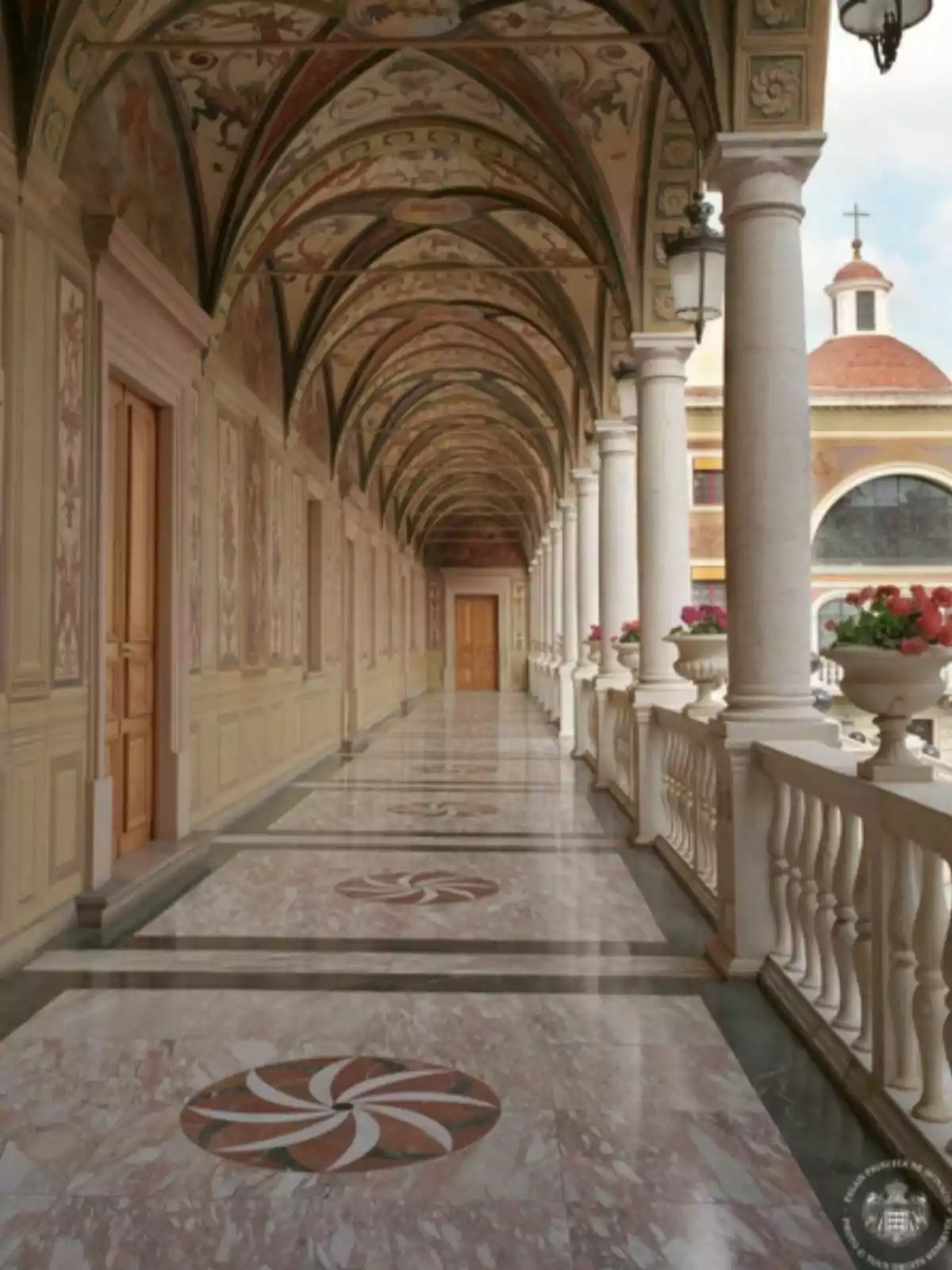 Pasillo con techo abovedado decorado con frescos, columnas blancas y piso de mármol con diseños circulares, al fondo se observa una iglesia con cúpula y cruz Pasillo con techo abovedado decorado con frescos, columnas blancas y piso de mármol con diseños circulares, al fondo se observa una iglesia con cúpula y cruz
