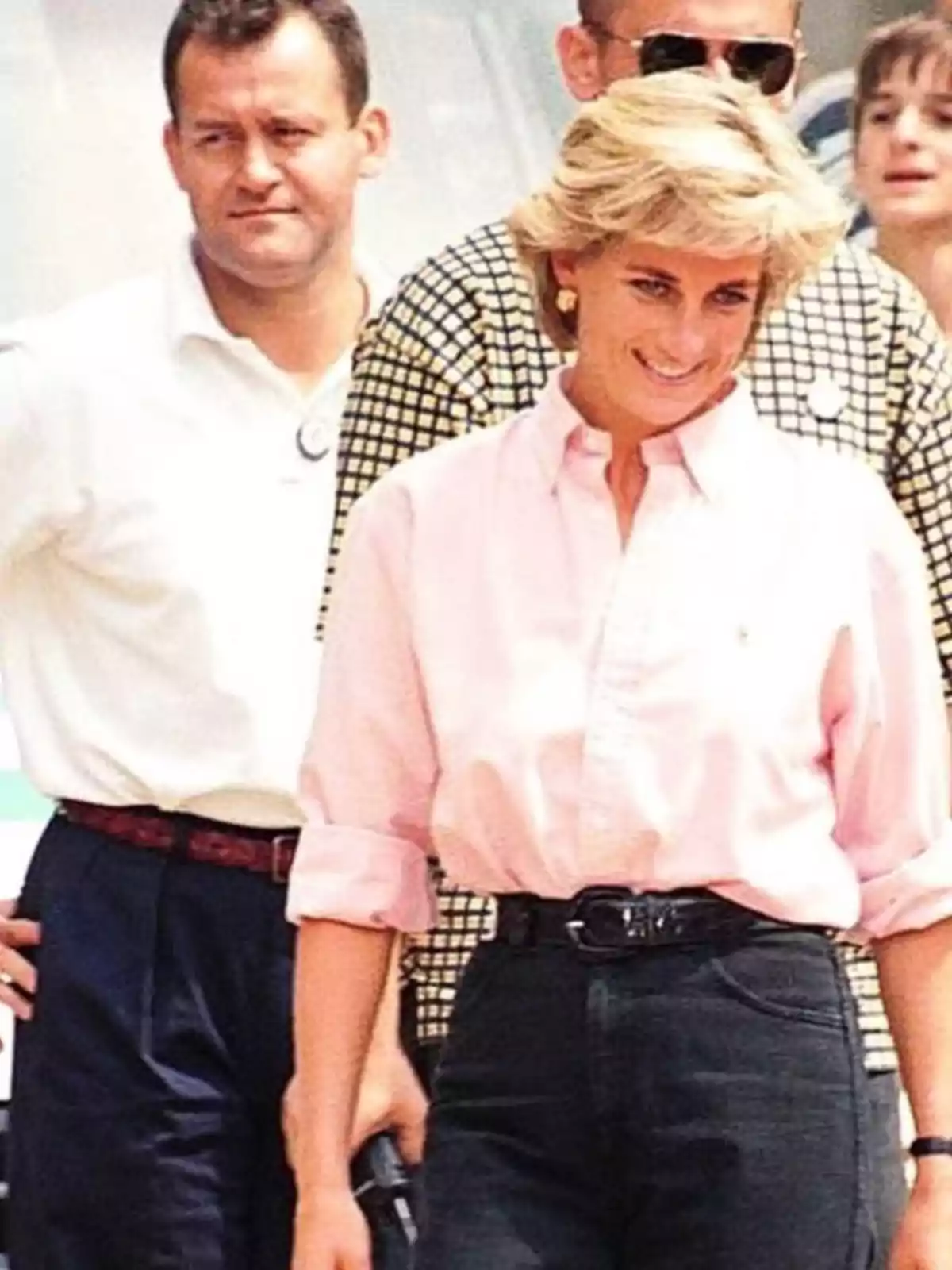 Mujer rubia con camisa rosa y pantalón oscuro sonríe mientras camina acompañada de varias personas en un ambiente al aire libre Mujer rubia con camisa rosa y pantalón oscuro sonríe mientras camina acompañada de varias personas en un ambiente al aire libre