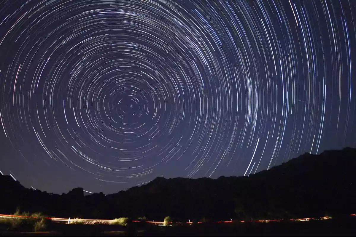 Cielo nocturno con estelas circulares de estrellas sobre silueta de montañas y luces de autos en la base Cielo nocturno con estelas circulares de estrellas sobre silueta de montañas y luces de autos en la base