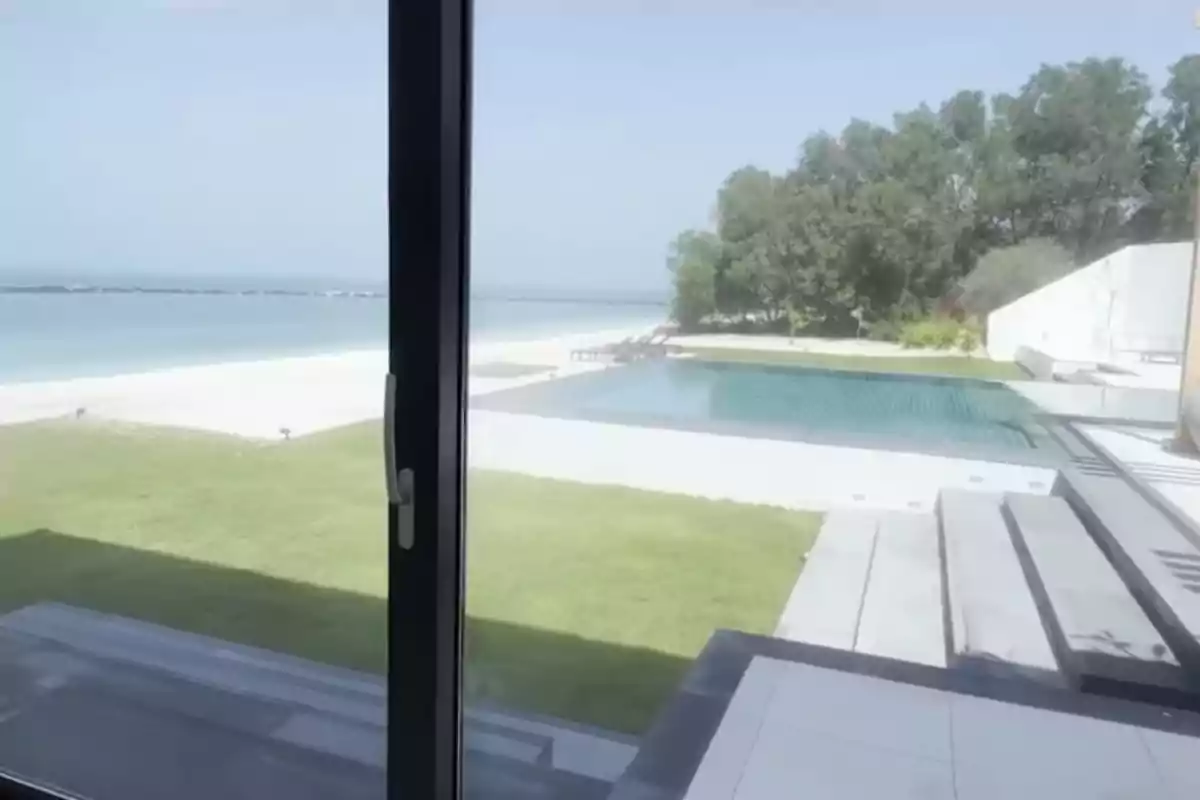 Vista desde una ventana hacia una piscina, césped, playa de arena blanca y el mar con árboles al fondo Vista desde una ventana hacia una piscina, césped, playa de arena blanca y el mar con árboles al fondo