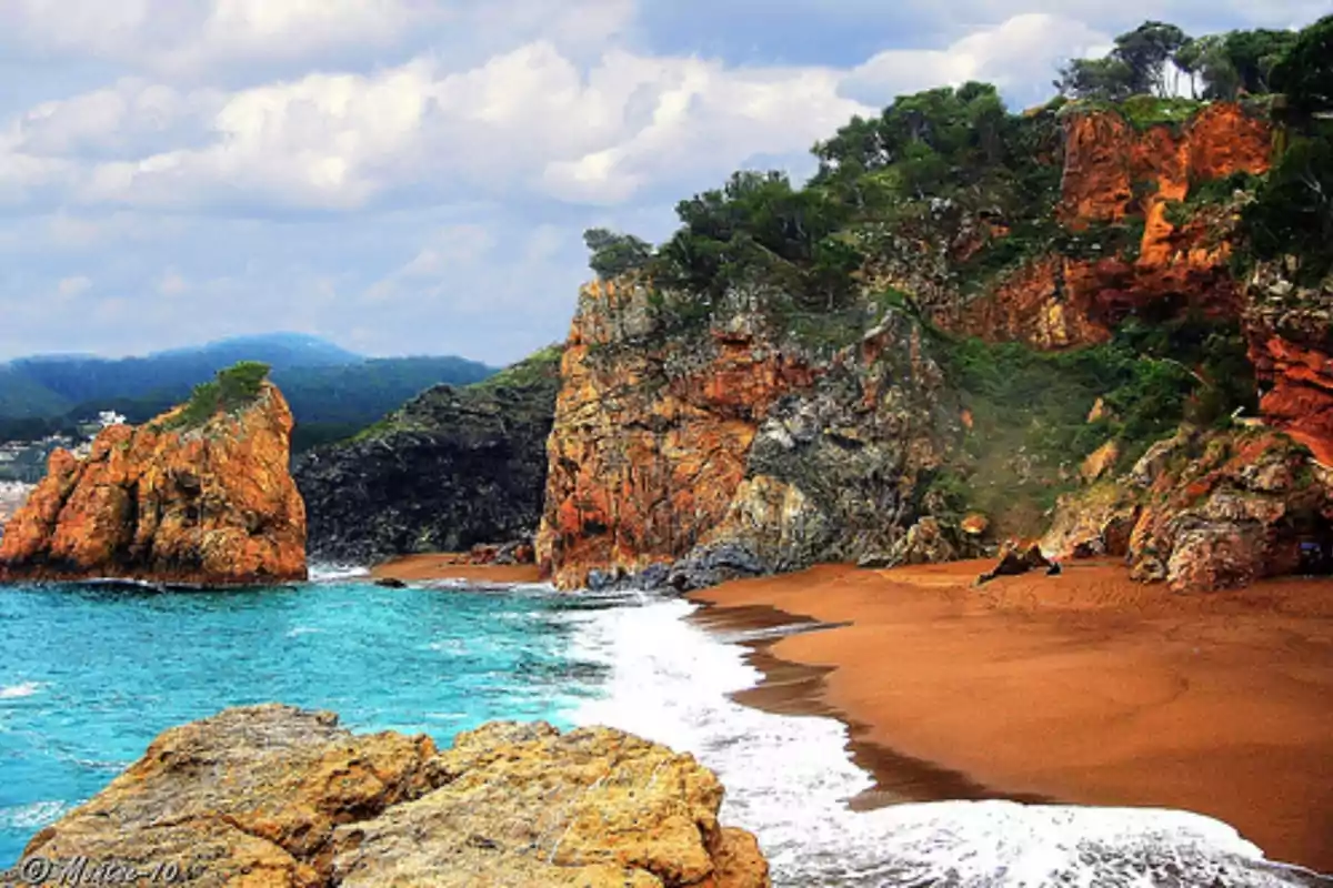 Platja de sorra daurada amb penya-segats rocosos i vegetació al costat d’un mar blau sota un cel ennuvolat Platja de sorra daurada amb penya-segats rocosos i vegetació al costat d’un mar blau sota un cel ennuvolat