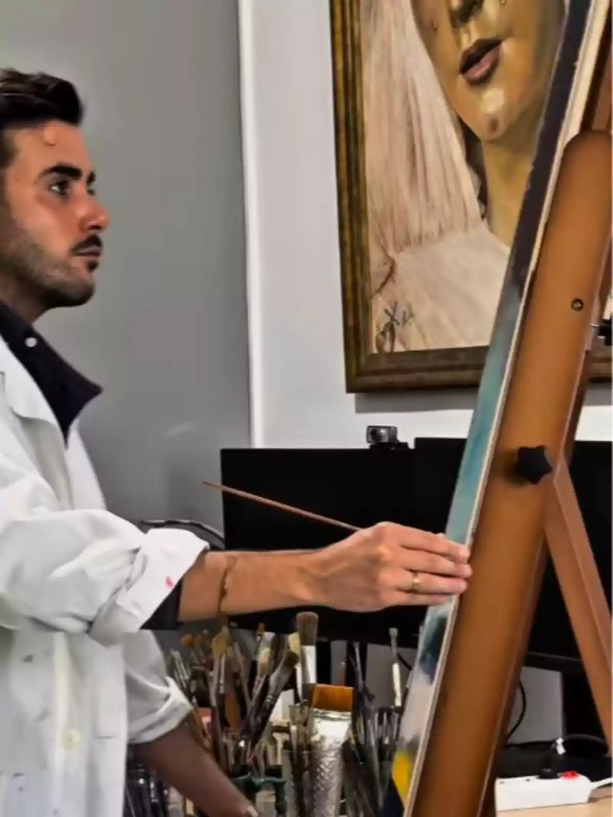 Alberto Rubio in a white coat painting at an easel in an art studio, surrounded by brushes and materials, with a painting of a woman in the background Alberto Rubio in a white coat painting at an easel in an art studio, surrounded by brushes and materials, with a painting of a woman in the background