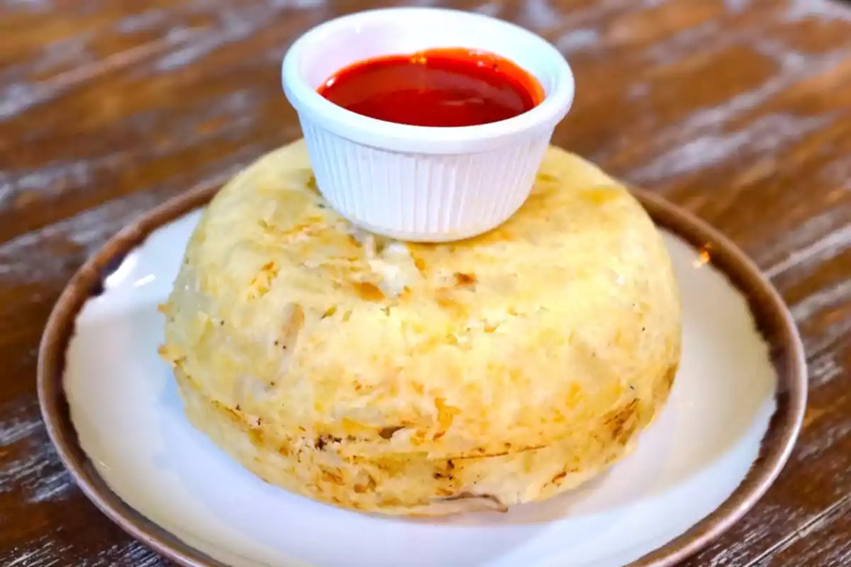 Plato de mofongo servido en un plato blanco con un recipiente pequeño de salsa roja encima Plato de mofongo servido en un plato blanco con un recipiente pequeño de salsa roja encima