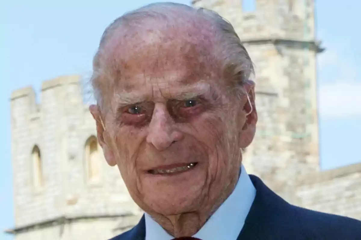 Philip of Edinburgh in a dark suit smiles in front of an ancient stone building.