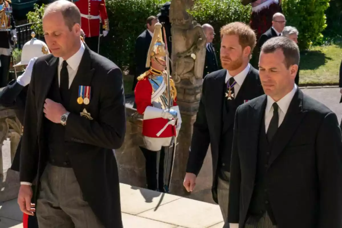 Los príncipes Guillermo y Harry y Peter Phillips vestidos de traje formal caminan juntos en un evento solemne al aire libre mientras un guardia con uniforme ceremonial se encuentra al fondo Los príncipes Guillermo y Harry y Peter Phillips vestidos de traje formal caminan juntos en un evento solemne al aire libre mientras un guardia con uniforme ceremonial se encuentra al fondo