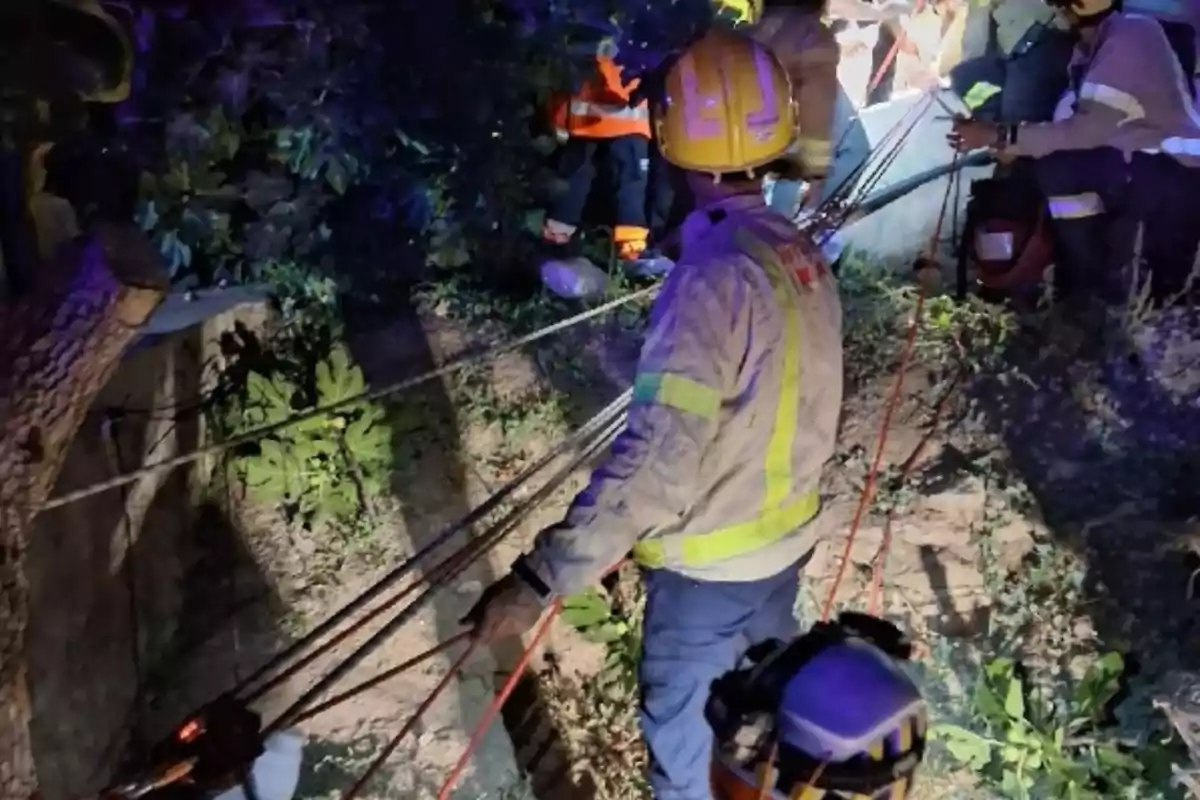 Diversos rescatadors amb cascos i equip de protecció treballen en una operació nocturna utilitzant cordes en una zona amb vegetació i terreny irregular. Diversos rescatadors amb cascos i equip de protecció treballen en una operació nocturna utilitzant cordes en una zona amb vegetació i terreny irregular.