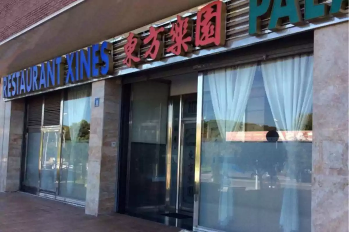 Façana d’un restaurant xinès amb rètol en lletres blaves i caràcters xinesos vermells, cortines blanques a les finestres i porta de vidre. Façana d’un restaurant xinès amb rètol en lletres blaves i caràcters xinesos vermells, cortines blanques a les finestres i porta de vidre.