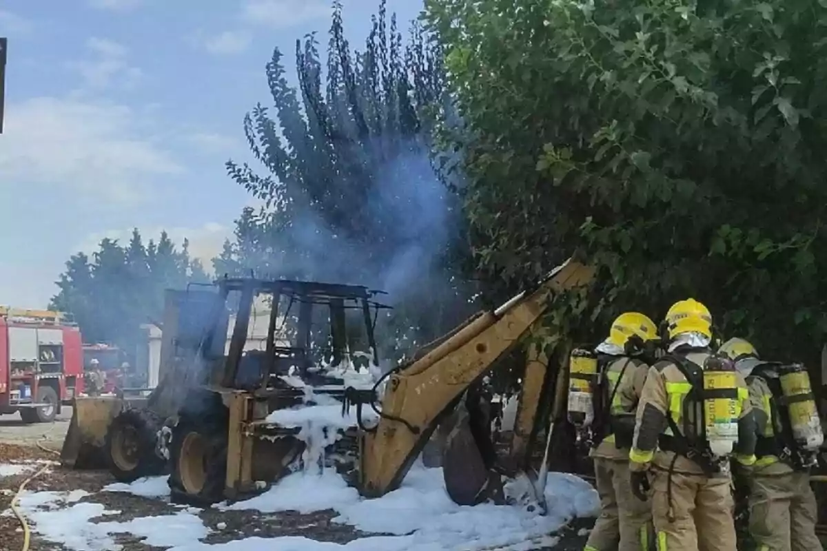 Tres bomberos con equipo de protección observan una retroexcavadora cubierta de espuma tras un incendio mientras una unidad de bomberos se encuentra al fondo