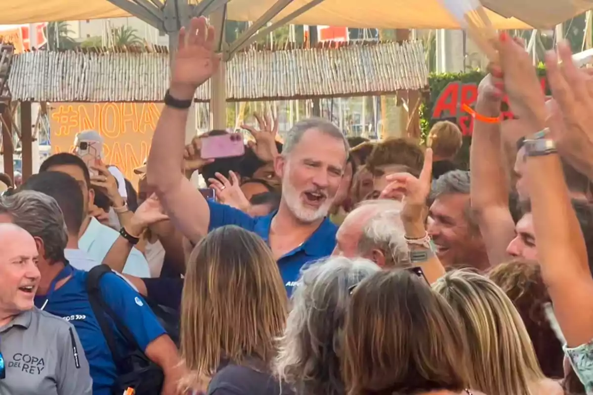 Un home somrient amb barba i samarreta blava aixeca la mà mentre està envoltat d’una multitud de persones que celebren i aplaudeixen en un ambient festiu a l’aire lliure. Un home somrient amb barba i samarreta blava aixeca la mà mentre està envoltat d’una multitud de persones que celebren i aplaudeixen en un ambient festiu a l’aire lliure.
