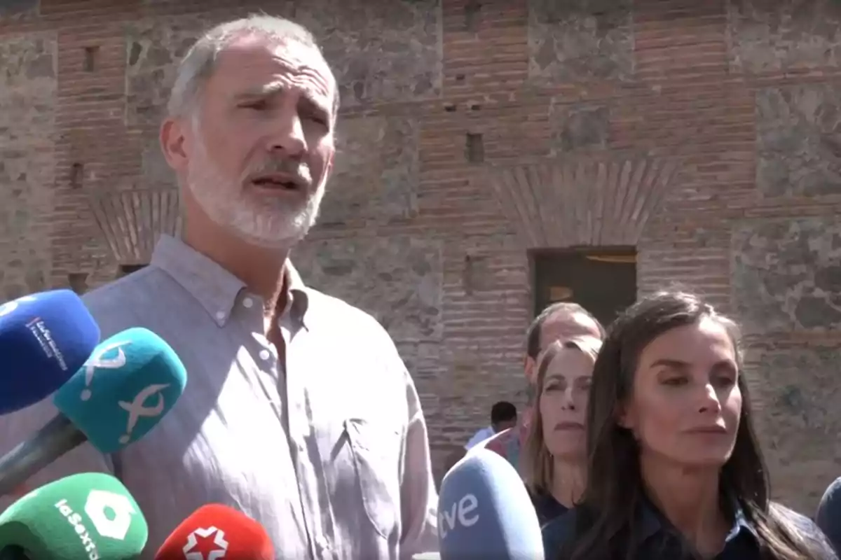 El rei Felip de cabells canosos i barba, juntament amb la reina Letícia de cabells llargs i foscos, són davant de diversos micròfons de diferents mitjans de comunicació en un entorn exterior amb una paret de maó i pedra de fons. El rei Felip de cabells canosos i barba, juntament amb la reina Letícia de cabells llargs i foscos, són davant de diversos micròfons de diferents mitjans de comunicació en un entorn exterior amb una paret de maó i pedra de fons.