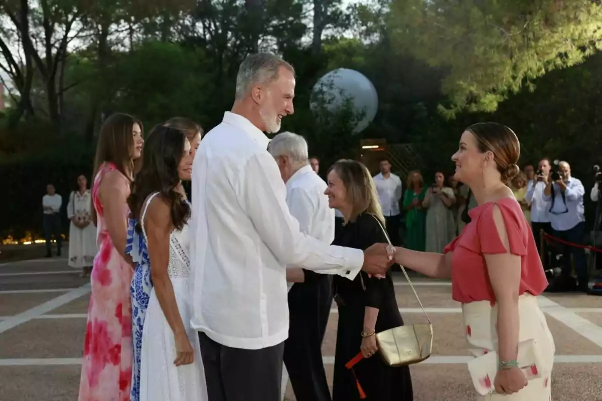 A tall man in a white jersey greets a woman dressed in light pink at an outdoor event surrounded by other elegantly dressed people and photographers in the background. A tall man in a white jersey greets a woman dressed in light pink at an outdoor event surrounded by other elegantly dressed people and photographers in the background.