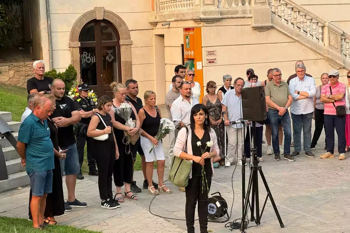 Un grup de persones reunides al carrer davant d’un edifici, algunes sostenen flors blanques i semblen estar participant en un acte solemne o commemoratiu. Un grup de persones reunides al carrer davant d’un edifici, algunes sostenen flors blanques i semblen estar participant en un acte solemne o commemoratiu.