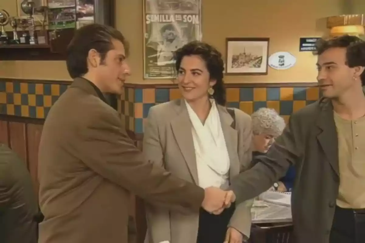 Tres personas vestidas de manera formal se saludan con un apretón de manos en el interior de una cafetería mientras una mujer mayor está sentada al fondo Tres personas vestidas de manera formal se saludan con un apretón de manos en el interior de una cafetería mientras una mujer mayor está sentada al fondo