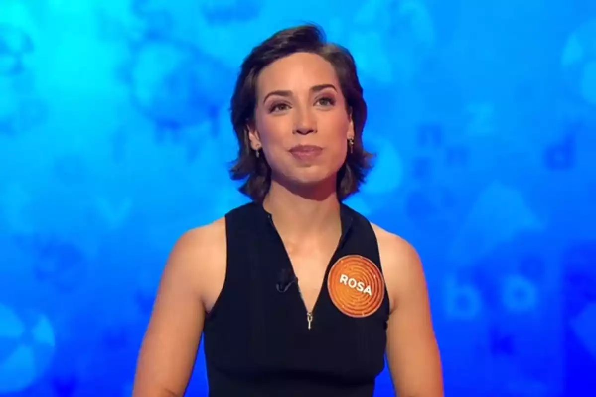 Rosa Rodríguez de cabello corto y oscuro con un vestido negro y una etiqueta naranja con el nombre Rosa sobre un fondo azul Rosa Rodríguez de cabello corto y oscuro con un vestido negro y una etiqueta naranja con el nombre Rosa sobre un fondo azul