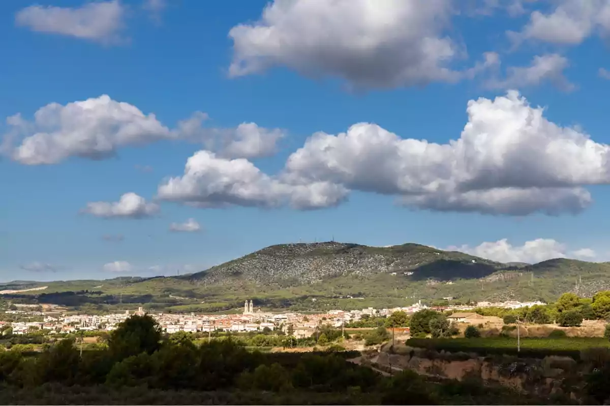 Vista panoràmica d’un poble envoltat de turons verds sota un cel blau amb núvols esponjosos Vista panoràmica d’un poble envoltat de turons verds sota un cel blau amb núvols esponjosos