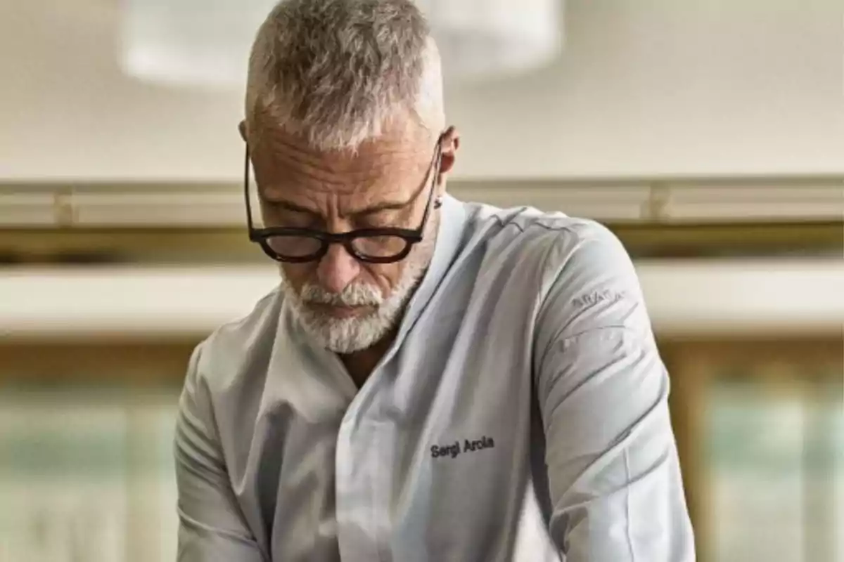 Hombre de cabello y barba canosos con gafas y chaqueta de chef trabajando en una cocina