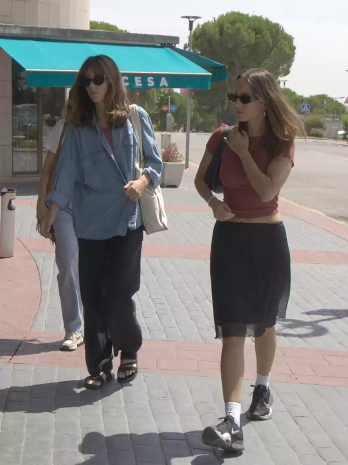 Silvia Alonso camina con otra mujer por una acera en un día soleado, ambas llevan gafas de sol y ropa casual, una viste camisa azul y pantalón negro, la otra camiseta roja y falda negra, al fondo se ve una marquesina verde y árboles Silvia Alonso camina con otra mujer por una acera en un día soleado, ambas llevan gafas de sol y ropa casual, una viste camisa azul y pantalón negro, la otra camiseta roja y falda negra, al fondo se ve una marquesina verde y árboles