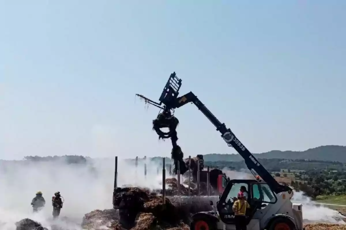 Un tractor amb braç mecànic aixeca bales de fenc mentre bombers treballen entre fum en un camp obert. Un tractor amb braç mecànic aixeca bales de fenc mentre bombers treballen entre fum en un camp obert.