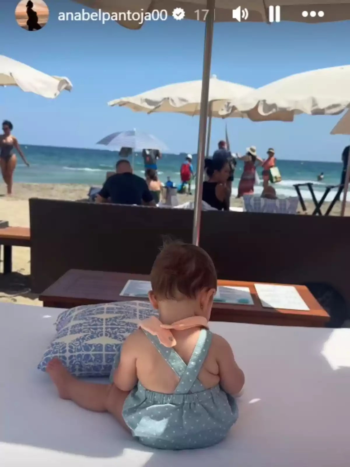 Story de Anabel Pantoja donde se ve a su hija Alma, sentada de espaldas sobre una cama en la playa bajo una sombrilla con el mar y varias personas al fondo Story de Anabel Pantoja donde se ve a su hija Alma, sentada de espaldas sobre una cama en la playa bajo una sombrilla con el mar y varias personas al fondo