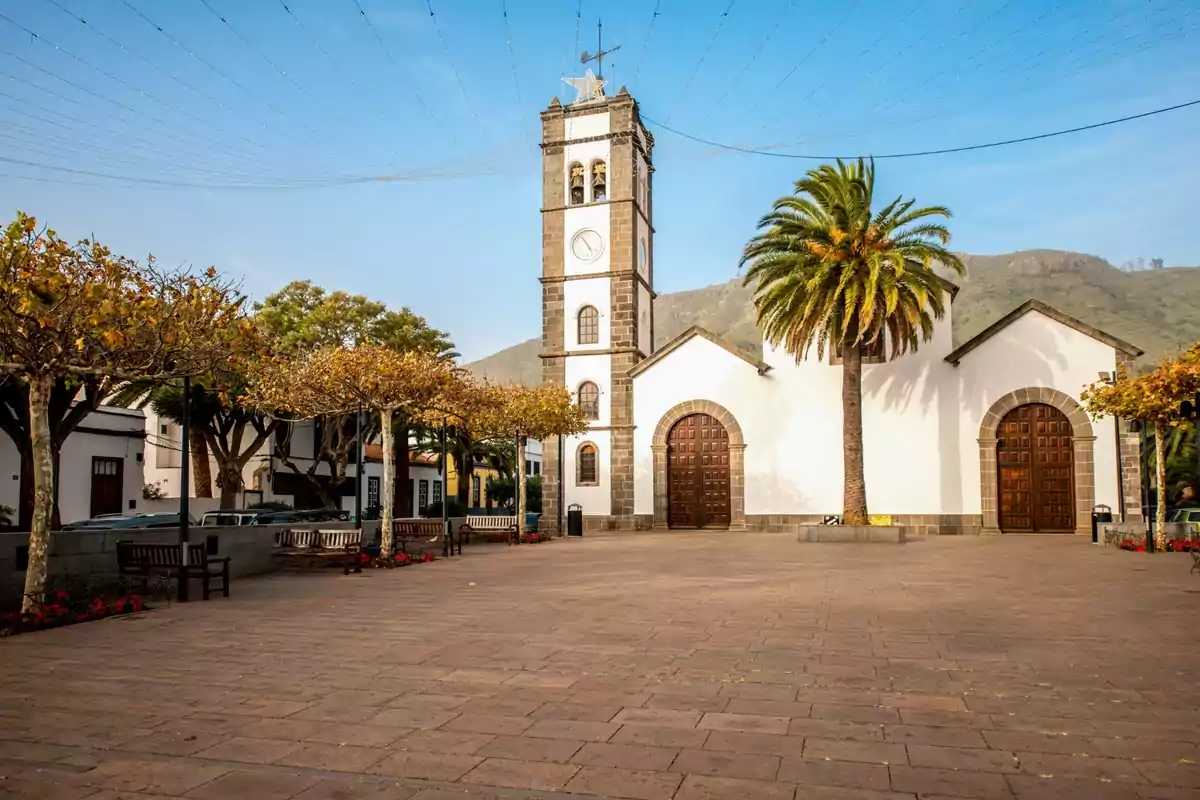 Plaza con bancos y árboles frente a una iglesia blanca con torre de reloj y una palmera en el centro Plaza con bancos y árboles frente a una iglesia blanca con torre de reloj y una palmera en el centro