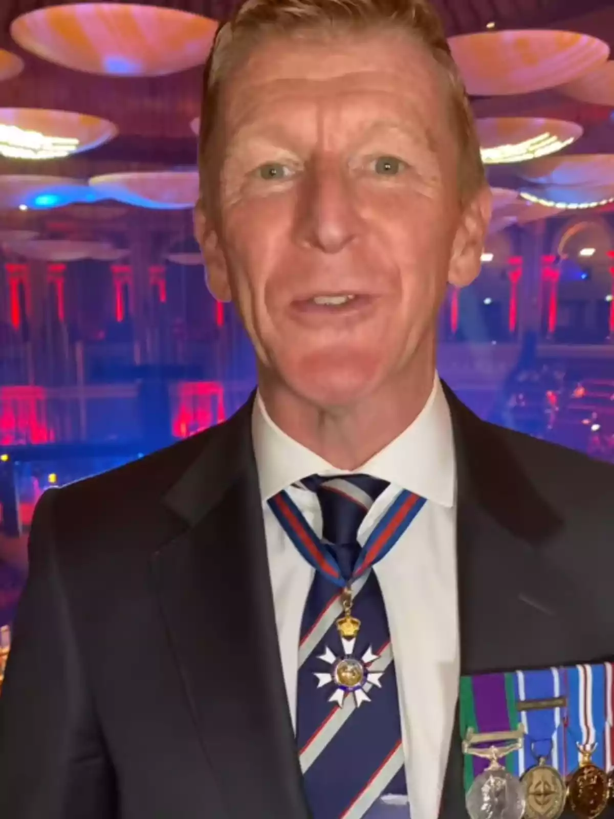 Home de cabell curt i ros vestit amb un vestit fosc i corbata, portant diverses medalles i amb un fons il·luminat en tons vermells i blaus Home de cabell curt i ros vestit amb un vestit fosc i corbata, portant diverses medalles i amb un fons il·luminat en tons vermells i blaus