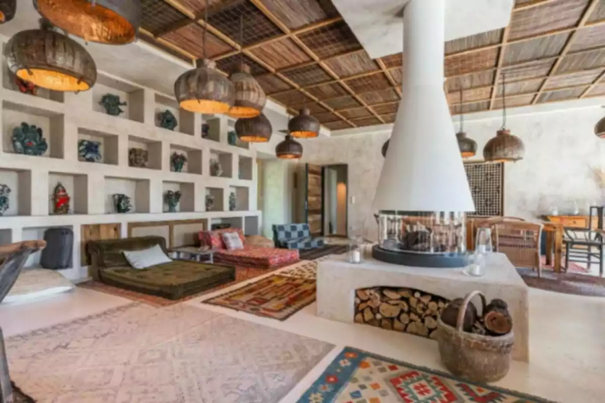 Sala de estilo bohemio con chimenea central, alfombras coloridas, lámparas colgantes de mimbre y estanterías empotradas decoradas con jarrones y objetos artesanales Sala de estilo bohemio con chimenea central, alfombras coloridas, lámparas colgantes de mimbre y estanterías empotradas decoradas con jarrones y objetos artesanales