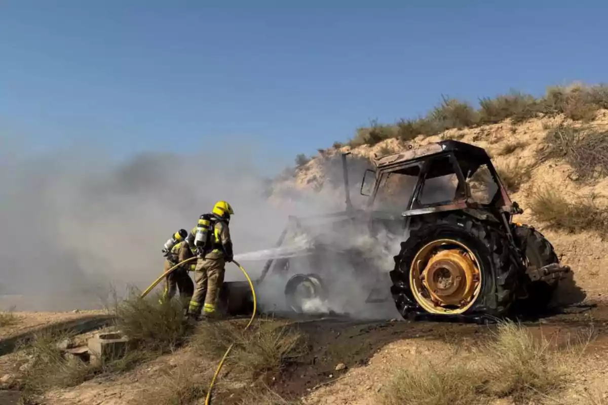 Bombers apaguen l’incendi d’un tractor en una zona rural amb vegetació seca i molta fum.