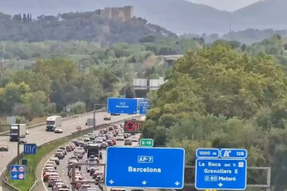Trànsit dens en una autopista envoltada d’arbres amb senyals blaves que indiquen la direcció cap a Barcelona i altres localitats.