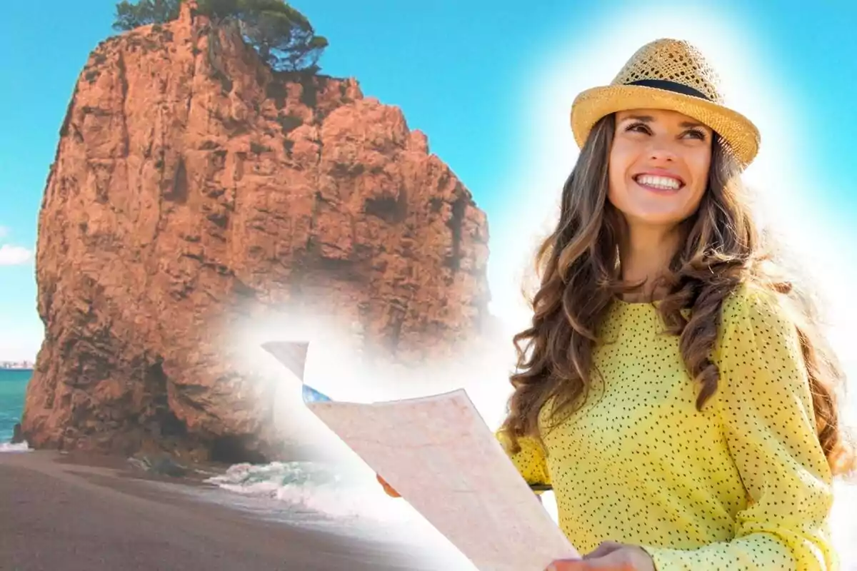 Dona somrient amb barret i brusa groga sosté un mapa davant d’una gran roca a la platja sota un cel blau