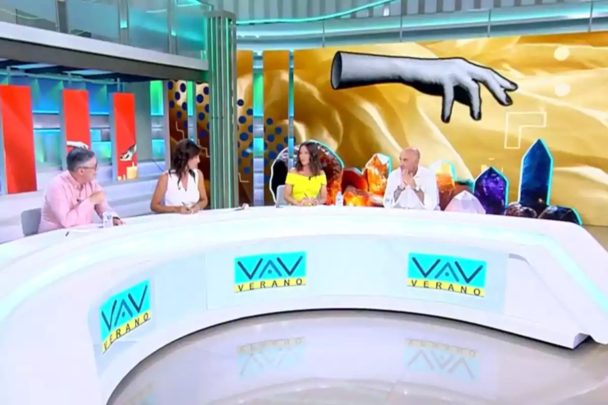 Cuatro personas conversan en un set de televisión moderno con una mesa blanca curva y una pantalla de fondo que muestra una mano ilustrada y varios cristales de colores. Captura de 'Vamos a ver' Cuatro personas conversan en un set de televisión moderno con una mesa blanca curva y una pantalla de fondo que muestra una mano ilustrada y varios cristales de colores. Captura de 'Vamos a ver'