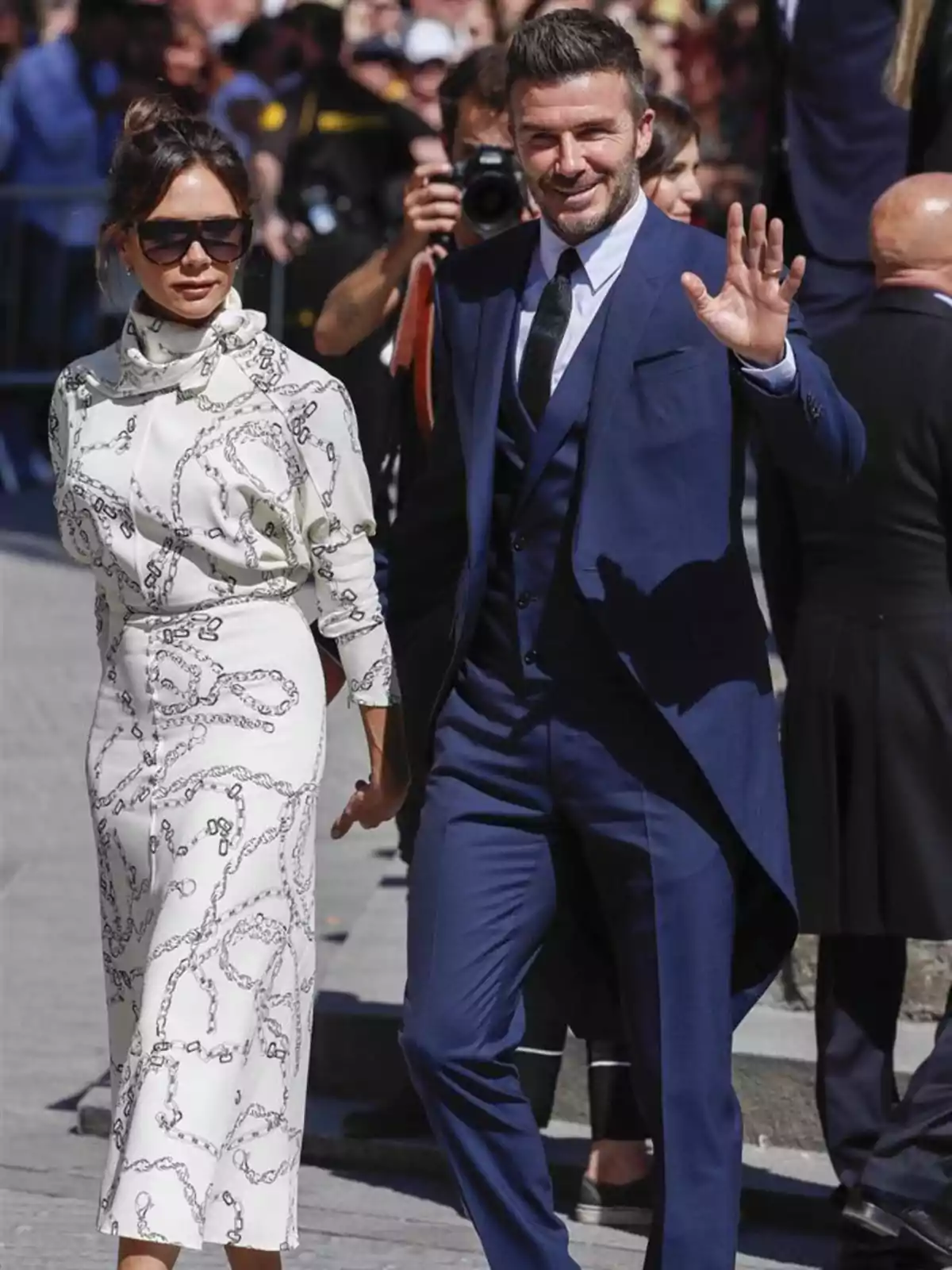 Una pareja elegantemente vestida camina de la mano en un evento al aire libre mientras él saluda sonriente y ella lleva gafas de sol Una pareja elegantemente vestida camina de la mano en un evento al aire libre mientras él saluda sonriente y ella lleva gafas de sol