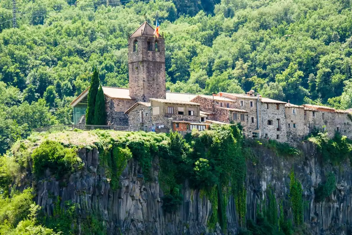 Poble de cases de pedra i una església amb torre, situat sobre un penya-segat envoltat de vegetació i bosc dens Poble de cases de pedra i una església amb torre, situat sobre un penya-segat envoltat de vegetació i bosc dens