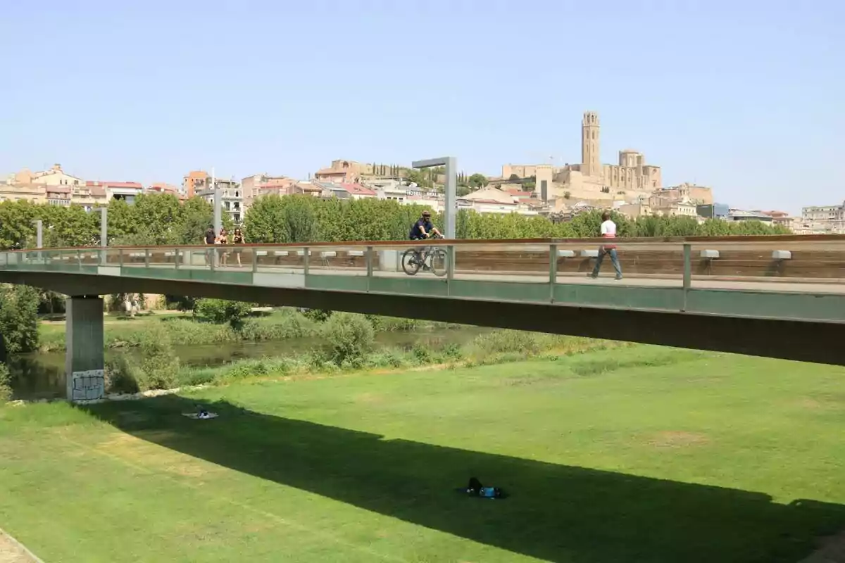 Puente peatonal con personas caminando y en bicicleta sobre un área verde con una ciudad y una catedral al fondo en un día soleado Puente peatonal con personas caminando y en bicicleta sobre un área verde con una ciudad y una catedral al fondo en un día soleado