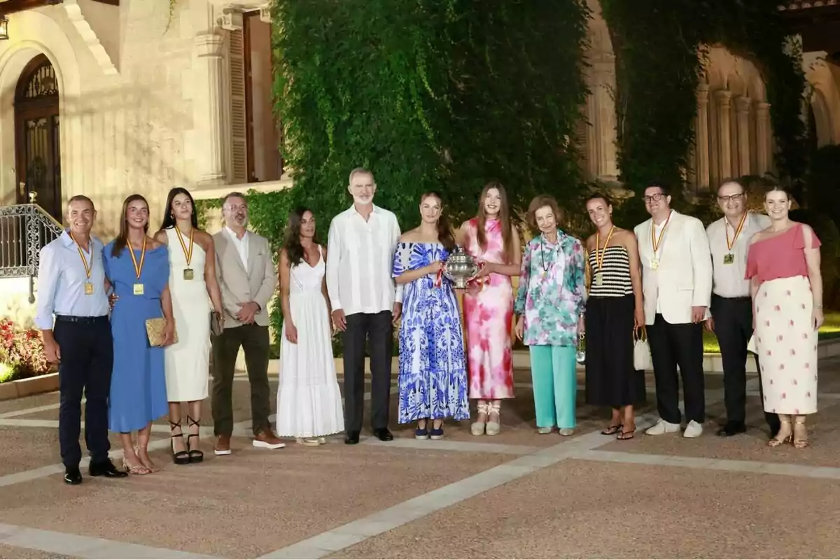 A group of elegantly dressed people poses outdoors in front of a building covered in greenery, some wearing medals and a woman holding a silver trophy. A group of elegantly dressed people poses outdoors in front of a building covered in greenery, some wearing medals and a woman holding a silver trophy.