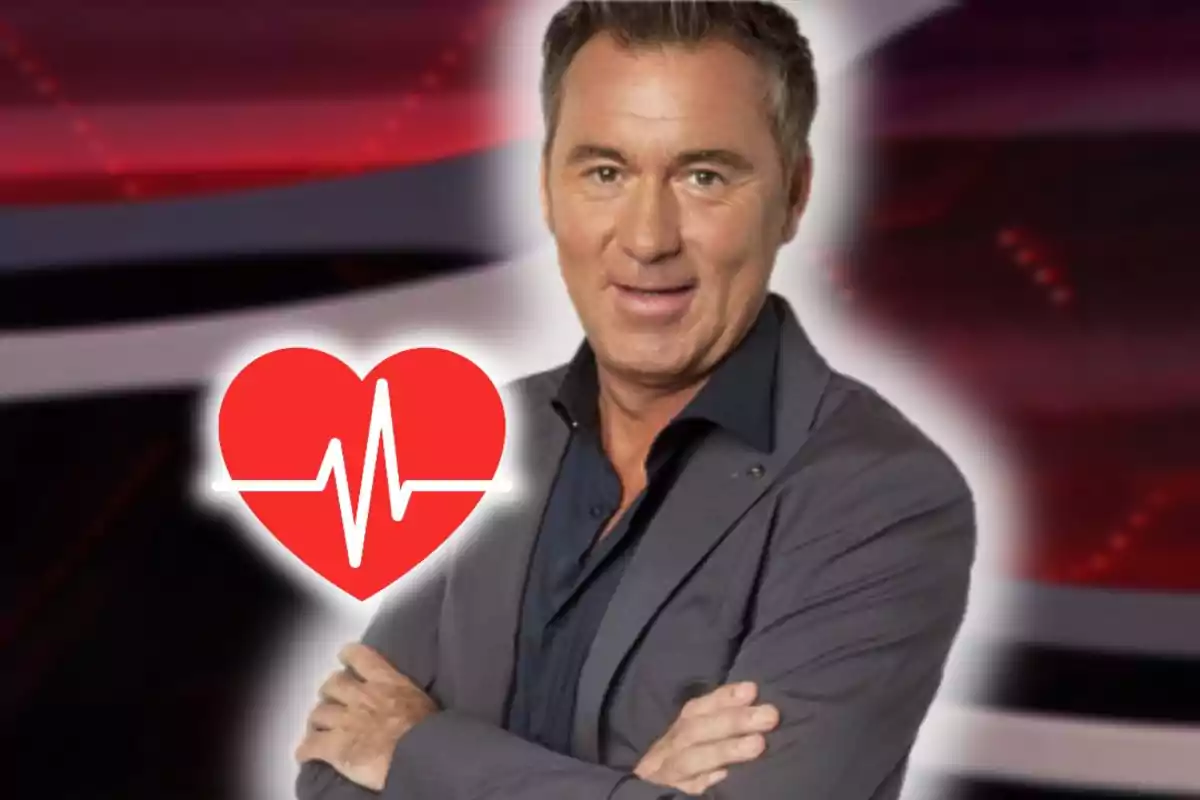 Home de cabell curt i canós amb americana grisa i camisa fosca somriu amb els braços creuats al costat d’una icona de cor vermell amb una línia d’electrocardiograma sobre un fons desenfocat.