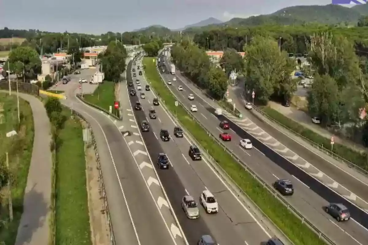 Vista aèria d’una autopista amb diversos carrils i nombrosos cotxes circulant en ambdues direccions, envoltada d’arbres i muntanyes al fons.