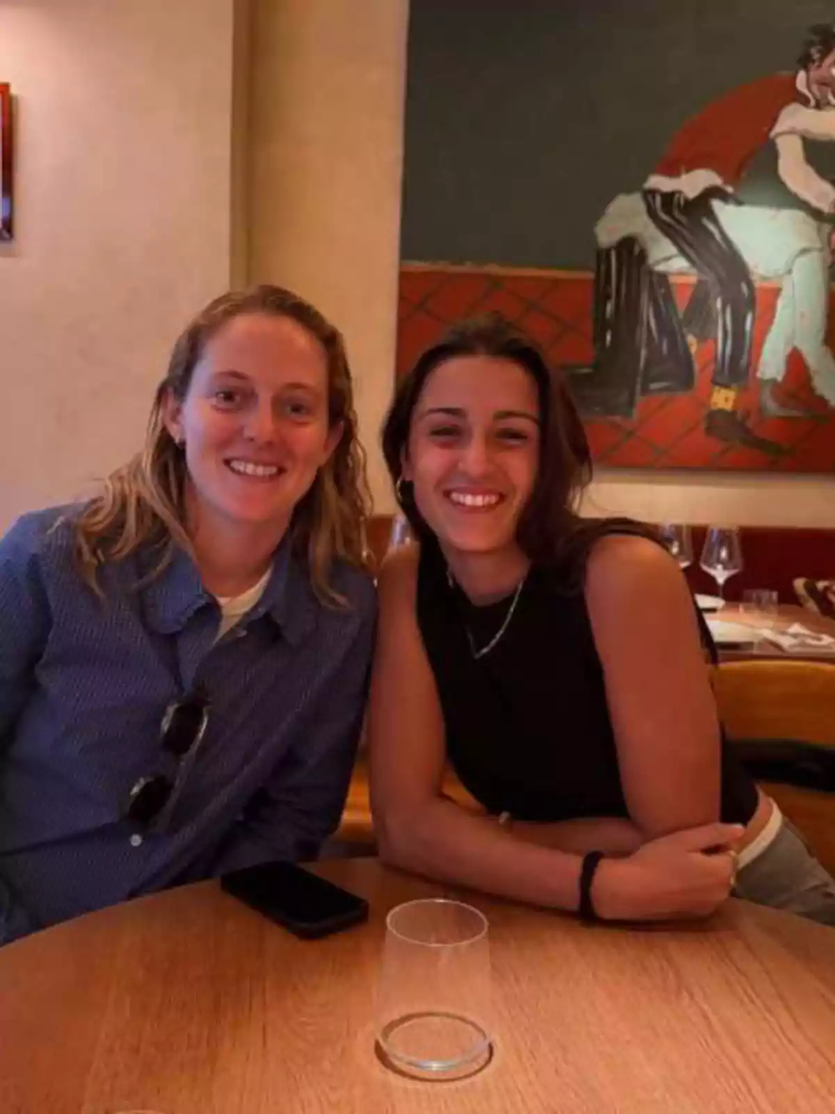 Dues dones somriuen assegudes en una taula de fusta en un restaurant amb una pintura colorida a la paret del fons. Dues dones somriuen assegudes en una taula de fusta en un restaurant amb una pintura colorida a la paret del fons.