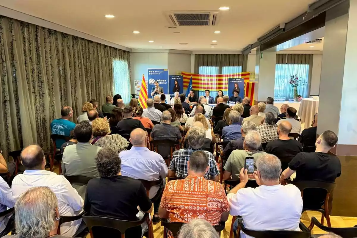 Un grup de persones assisteix a una conferència en una sala amb banderes catalanes i un panell de ponents al davant. Un grup de persones assisteix a una conferència en una sala amb banderes catalanes i un panell de ponents al davant.